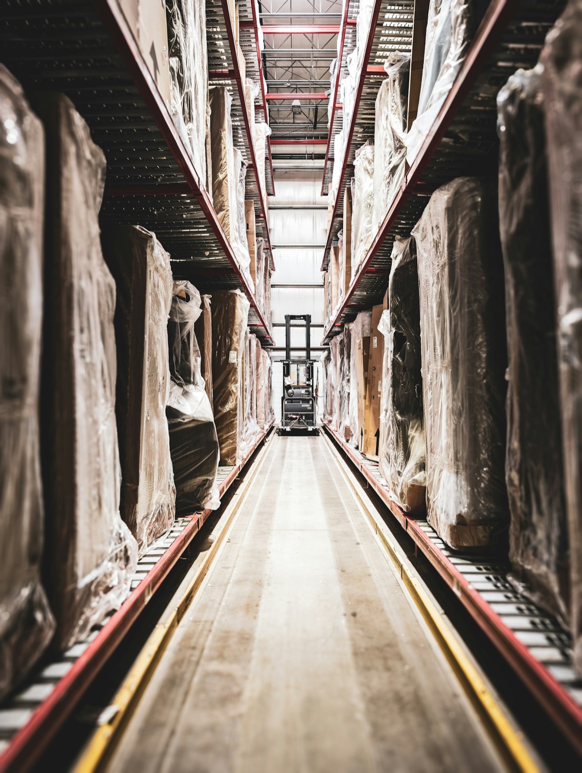 Warehouse aisle with wrapped pallets on tall racking and a forklift at the end